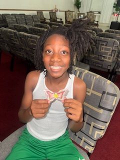 Smiling child with dreadlocks, wearing a white tank top and green shorts, sits on a patterned chair holding a handmade paper butterfly in a room with arranged chairs.