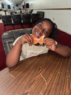 Smiling child sitting at a wooden table, holding a colorful butterfly craft with both hands, in a room with chairs and red carpet in the background.