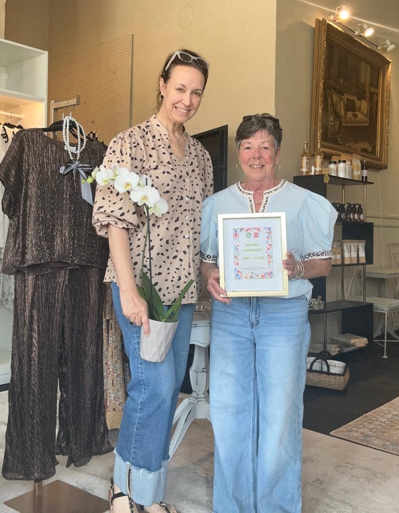 Two women smiling indoors; one holds a potted orchid, the other holds a framed certificate for National Garden Week. Clothing and decor items are visible in the background.