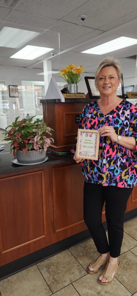 Woman standing at a counter holding a National Garden Week certificate, with a potted plant and yellow flowers in the background.