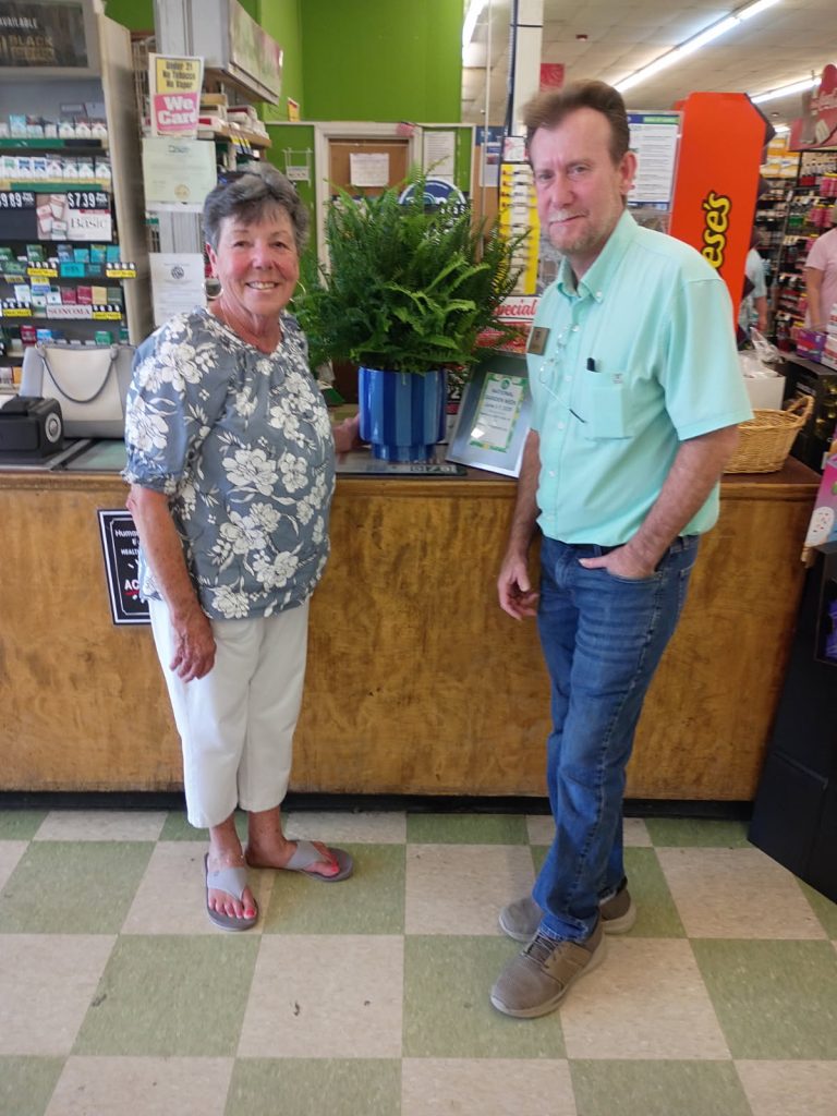 older woman and man stand smiling next to a green potted plant on a store counter. Store shelves and products are visible in the background.