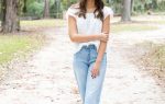 A woman stands on a tree-lined path outdoors, wearing a white top and light blue jeans, smiling with her arms crossed.
