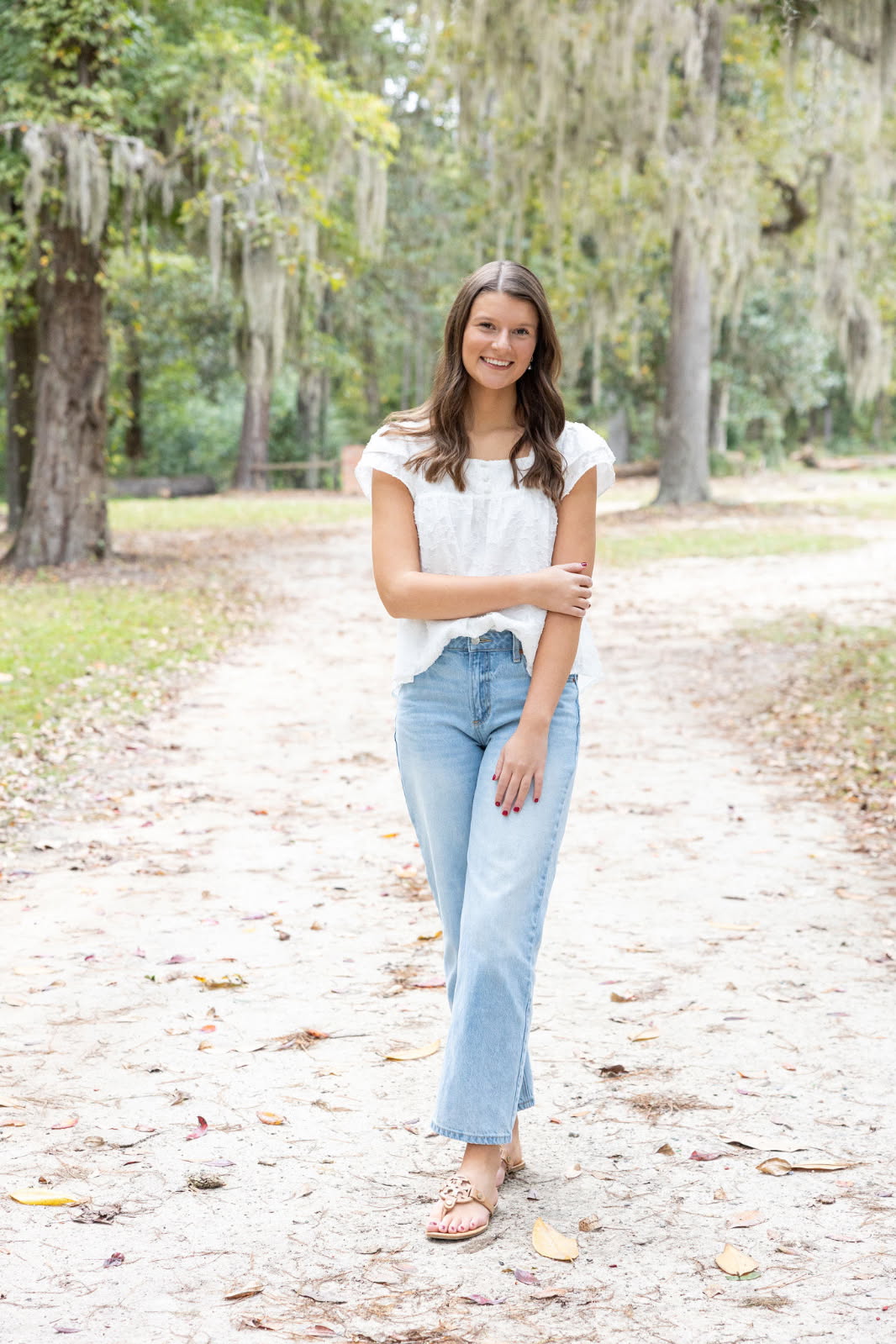 A woman stands on a tree-lined path outdoors, wearing a white top and light blue jeans, smiling with her arms crossed.