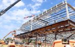 Steel framework of a building under construction, workers on lifts, crane, and flags flying at the top against a partly cloudy sky.