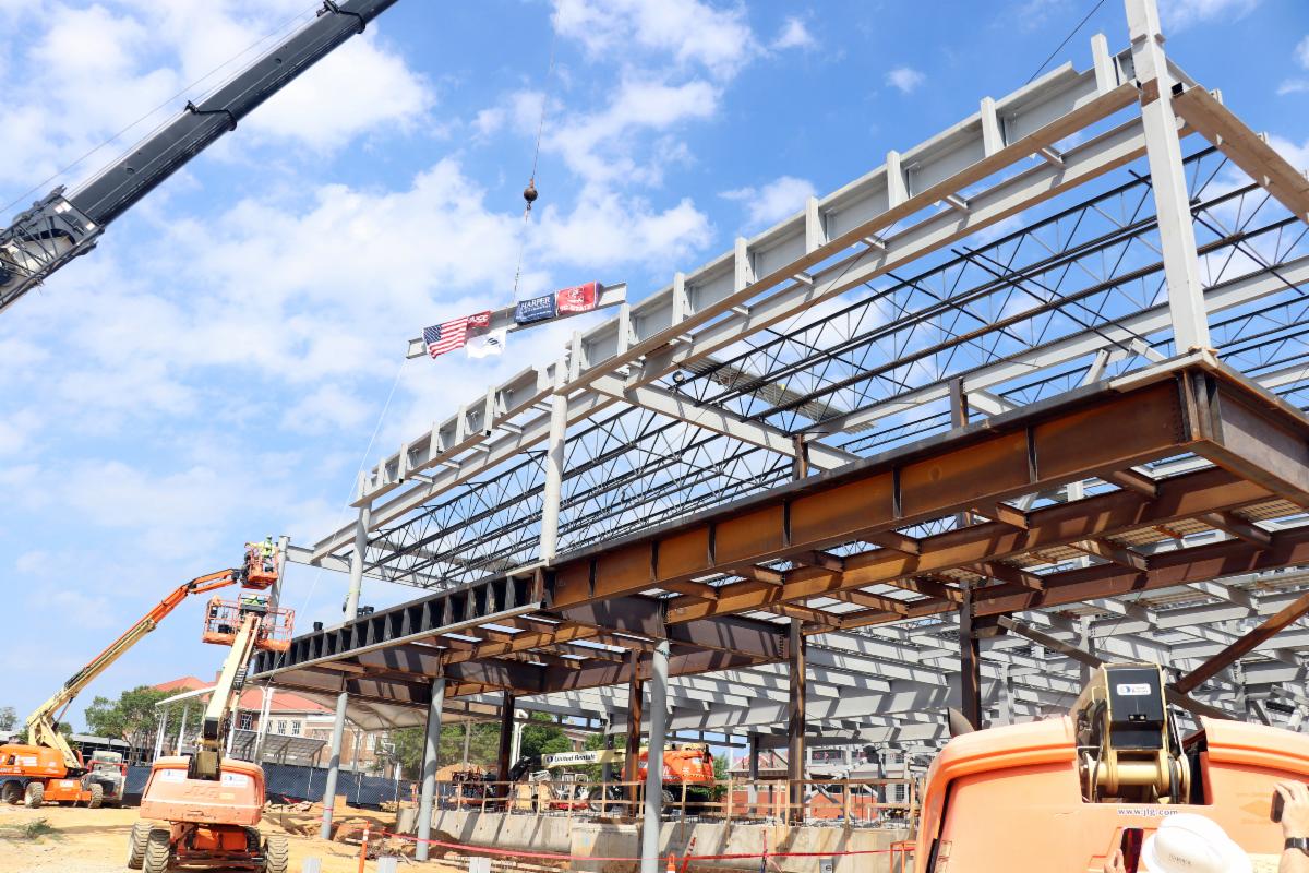 Steel framework of a building under construction, workers on lifts, crane, and flags flying at the top against a partly cloudy sky.