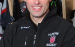 Man smiling in a black "Potsdam Hockey" jacket, standing in front of wooden lockers with sports equipment.