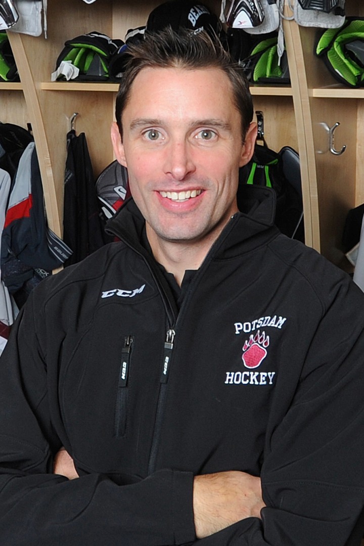 Man smiling in a black "Potsdam Hockey" jacket, standing in front of wooden lockers with sports equipment.