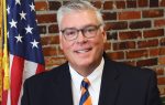Man in suit and striped tie smiling, wearing glasses, standing in front of a brick wall and an American flag.