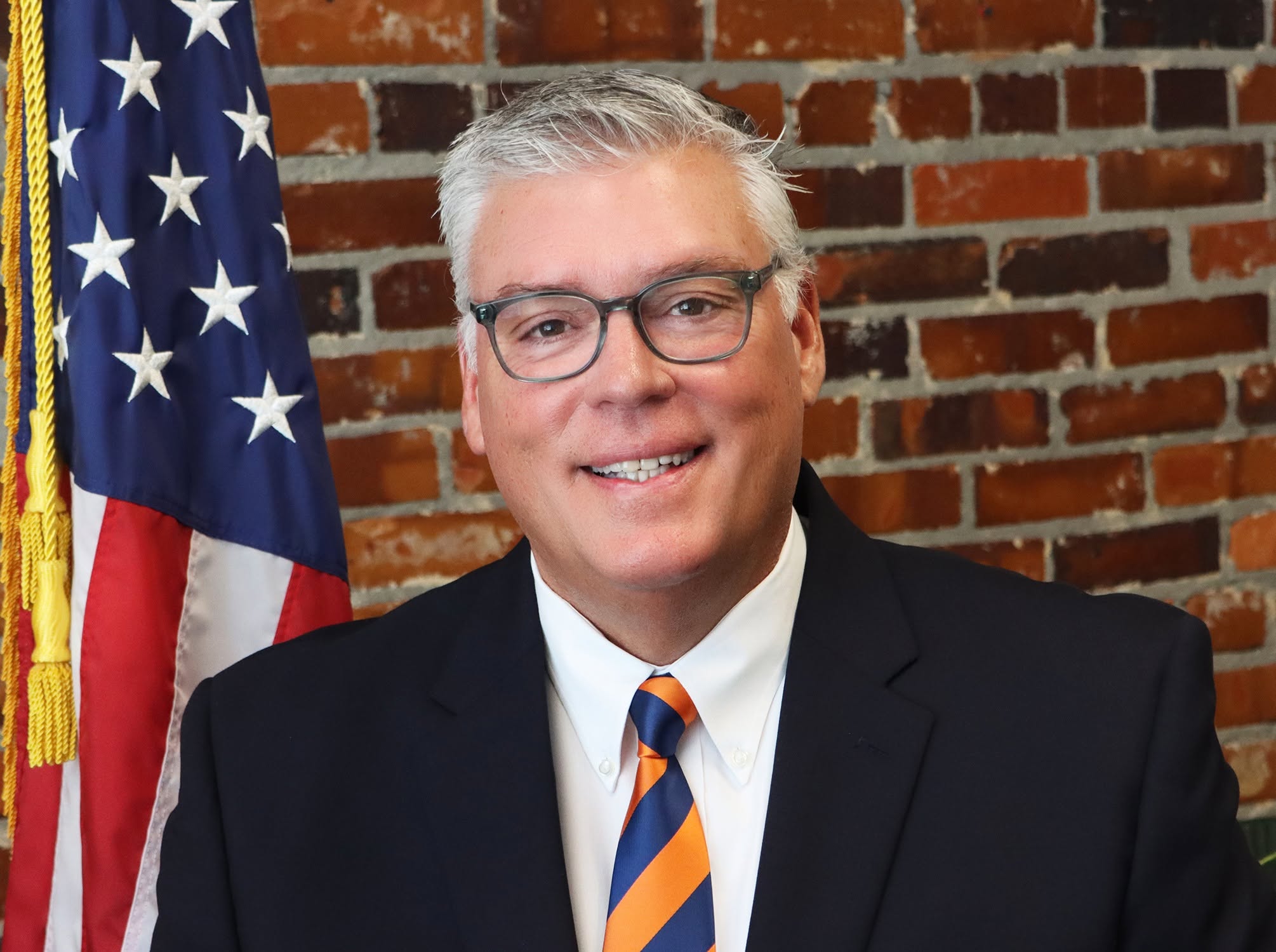 Man in suit and striped tie smiling, wearing glasses, standing in front of a brick wall and an American flag.