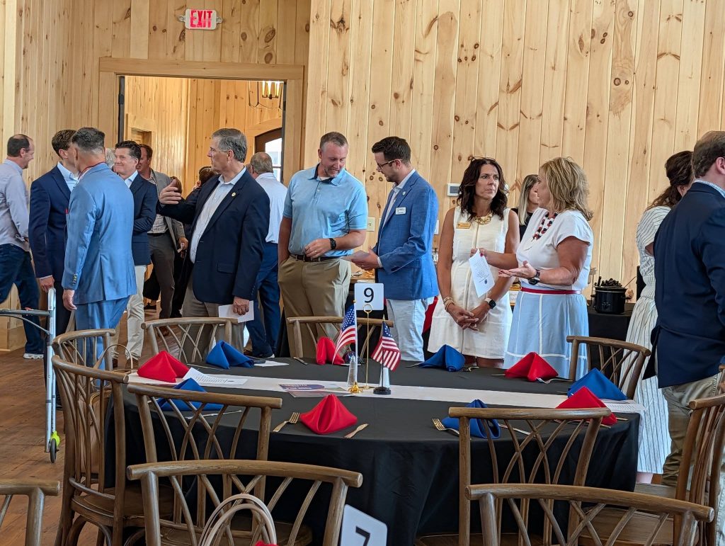 People in business attire converse around a round table set with red and blue napkins and small American flags in a wood-paneled event room.