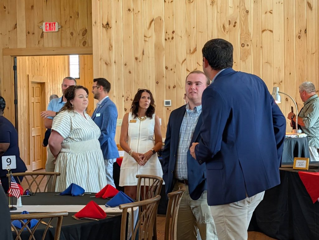 People in business attire stand and converse in a wooden banquet hall with decorated tables and red, white, and blue napkins.