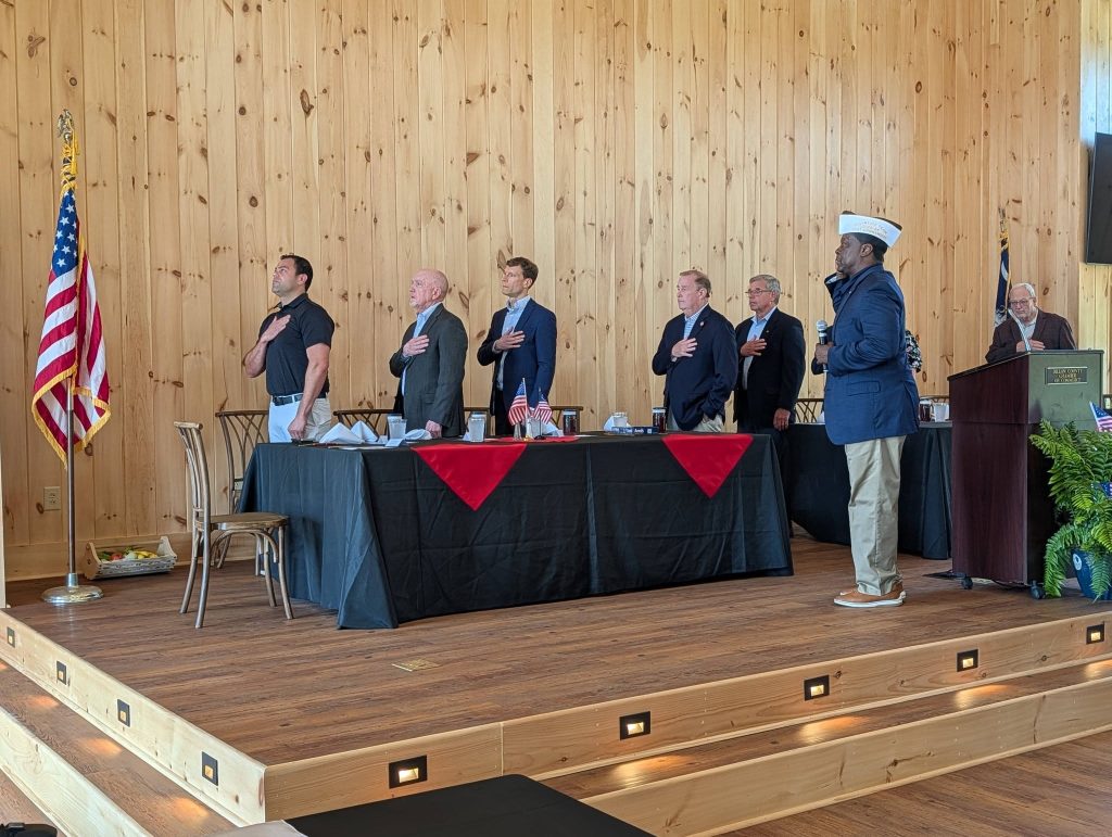 Seven men stand on a stage with hands over their hearts, facing the American flag, during what appears to be a formal or patriotic ceremony in a wooden hall.