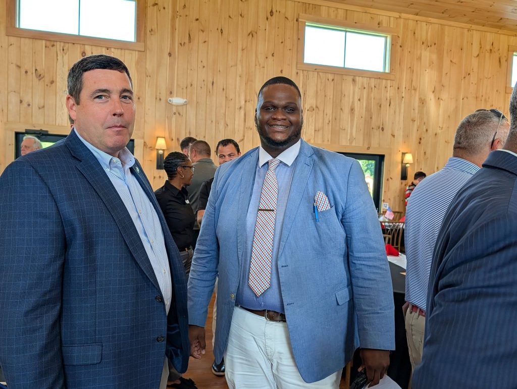 Two men in suits pose together at an indoor event with other people mingling in the background.
