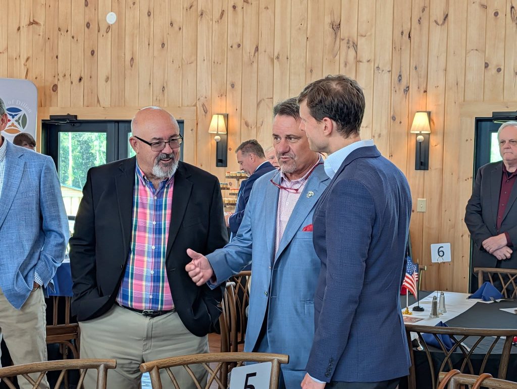 Three men in suits converse inside a wooden-walled room at a formal event, with tables, small American flags, and other attendees visible in the background.