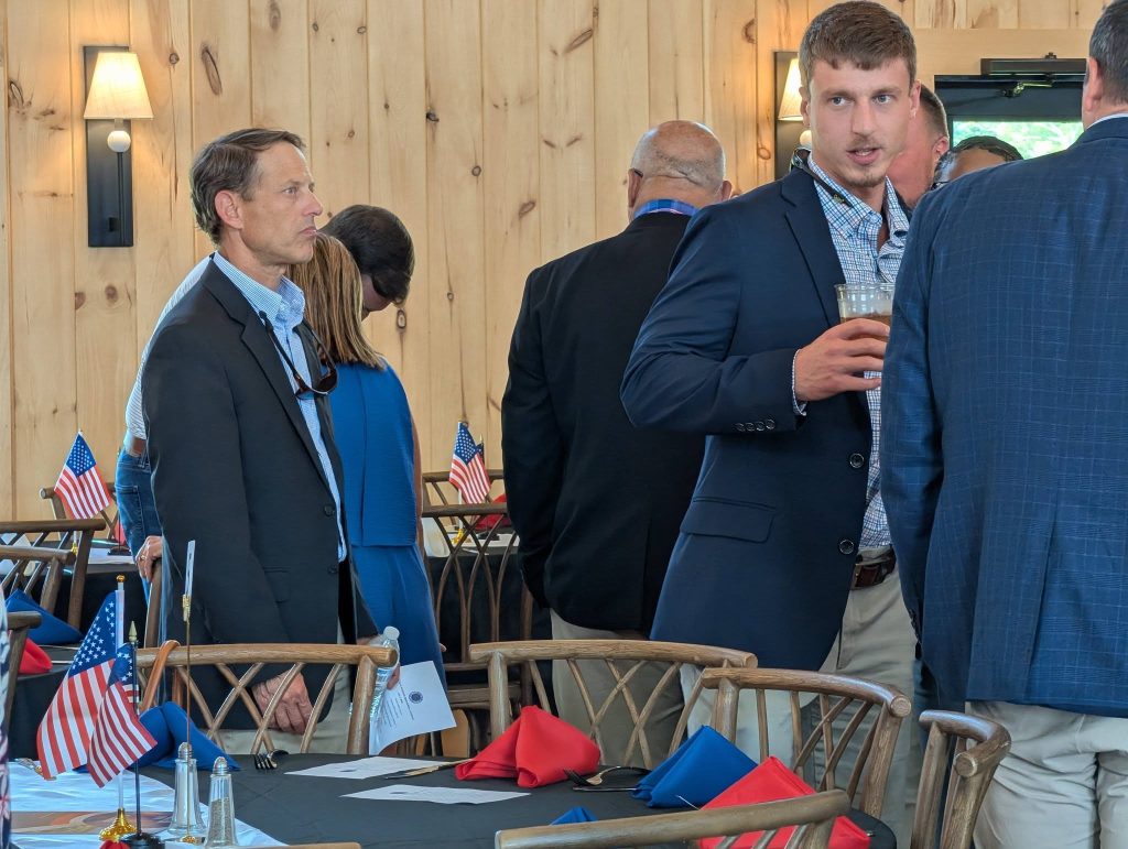 People in business attire at a formal indoor event, tables decorated with American flags, red and blue napkins, and wooden walls in the background.