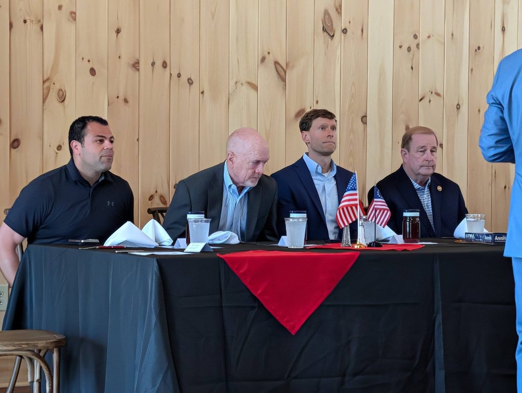 Four men sit at a table with small American flags, water glasses, and napkins in a wood-paneled room. One person stands off to the right.