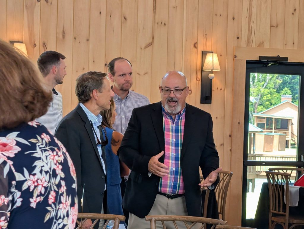 A group of people in business casual attire have a conversation inside a wooden-paneled room with natural light and outdoor view through a door.