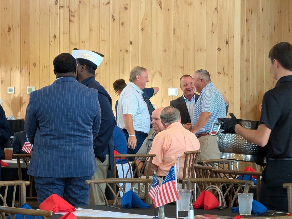 People socializing around tables decorated with American flags and colorful napkins in a wood-paneled room, with a server holding a metal ice bucket on the right.