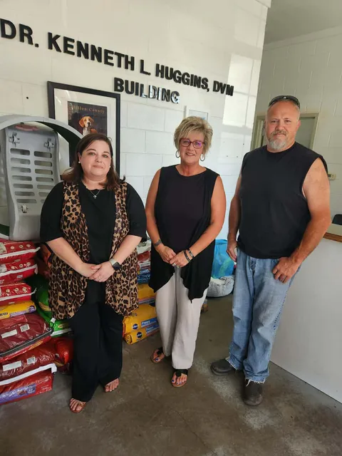Three adults stand in a veterinary clinic next to pet supplies, with a sign reading "Dr. Kenneth L. Huggins, DVM Building" on the wall behind them.