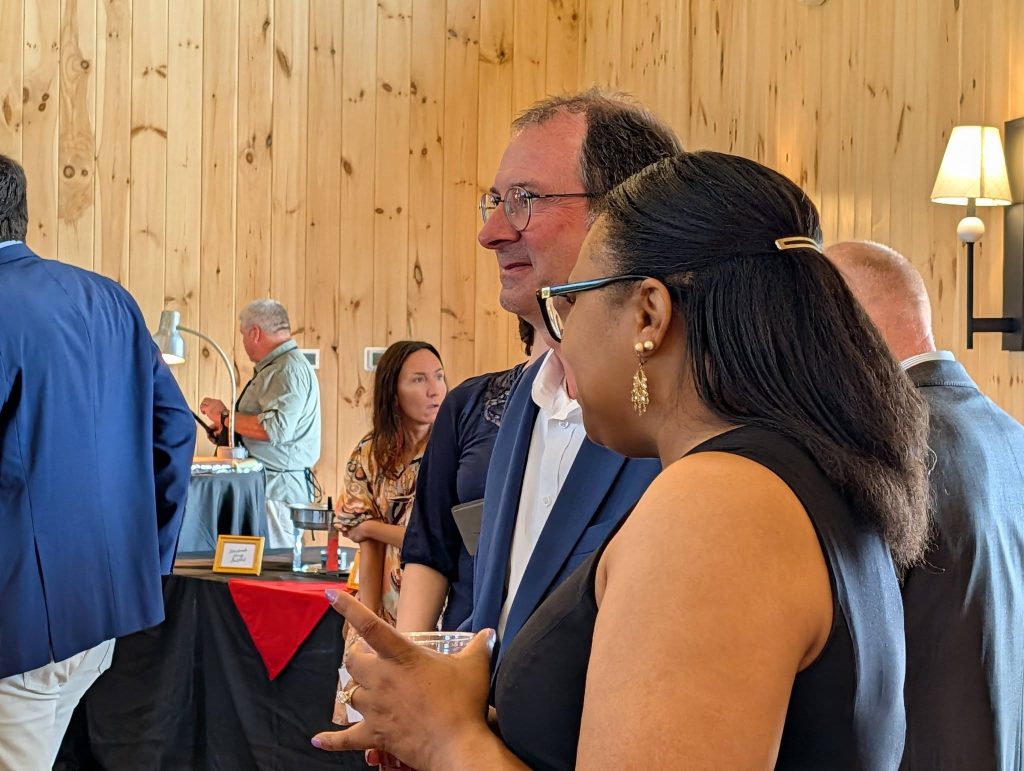 People socializing at a formal indoor event with wooden walls, some holding drinks, and a food station in the background.