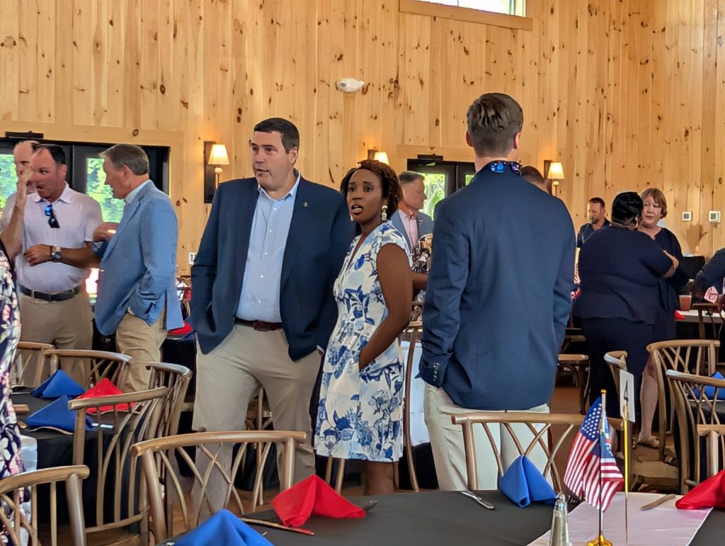 People conversing at an indoor event with wooden walls, dressed in business attire. Tables are set with red and blue napkins, and small American flags are displayed.