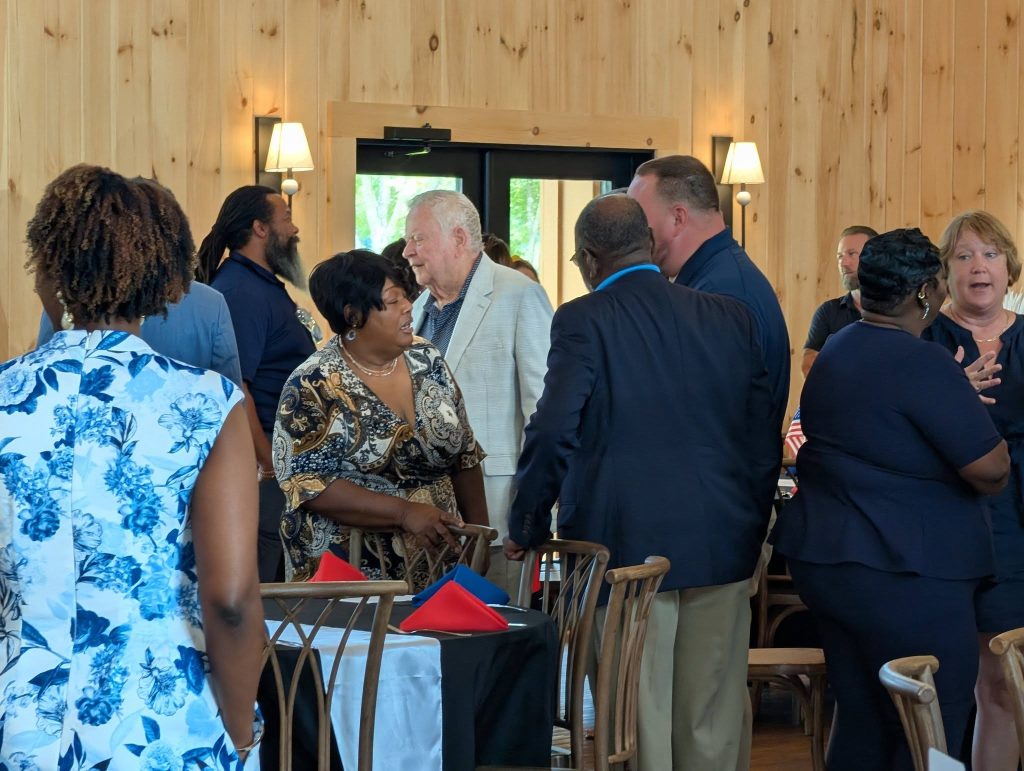 People standing and conversing in a warmly lit, wood-paneled room with tables and chairs, some dressed in business attire, appearing to be at a social or professional event.