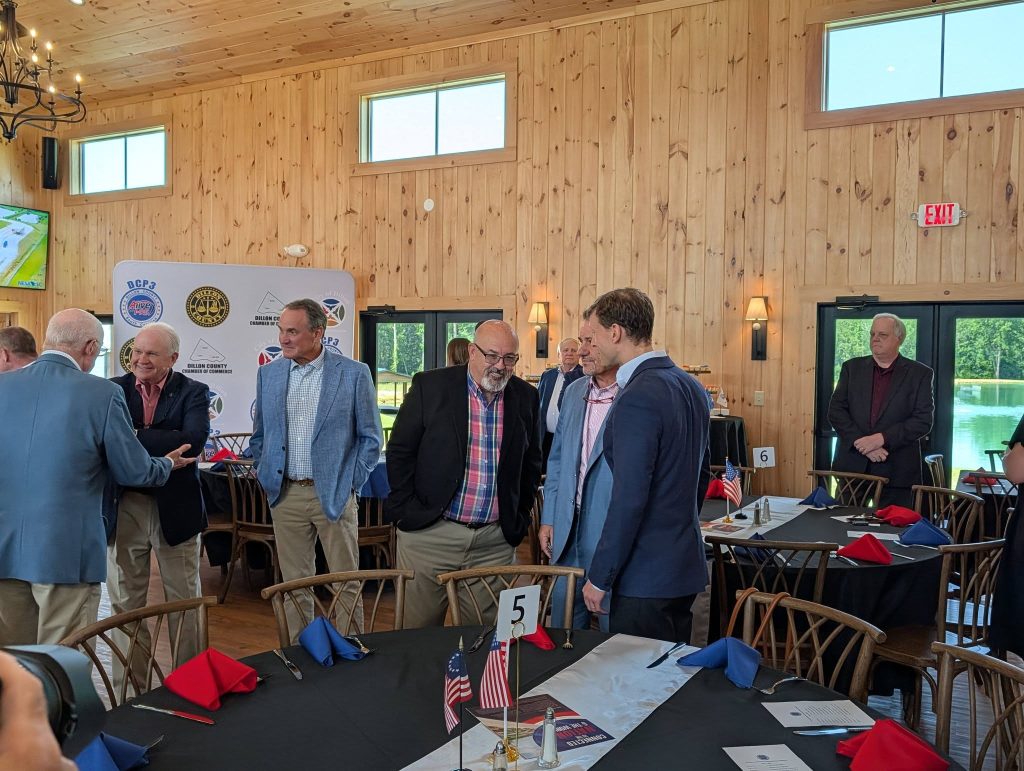 People dressed in business attire converse in a wooden banquet hall with decorated tables, American flags, and a branded backdrop in the background.