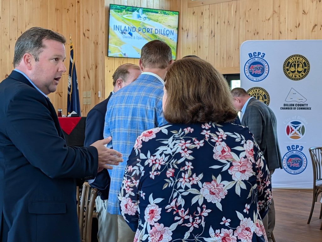 A group of people in business attire converse at an indoor event with banners and a screen displaying "Inland Port Dillon" in the background.