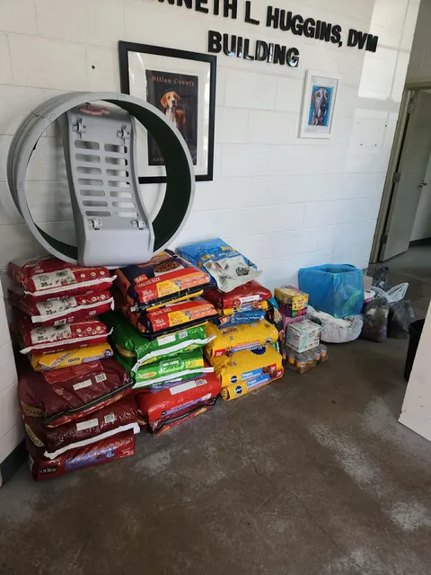 Bags of pet food and supplies stacked against a wall in a veterinary clinic, with artwork and a large exercise wheel nearby.
