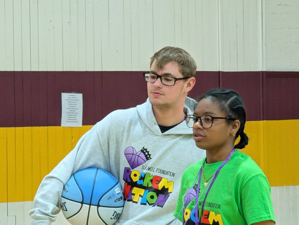 Two people in colorful shirts with basketball logos stand in a gym; one holds a blue-and-white basketball, both wear glasses and appear focused.