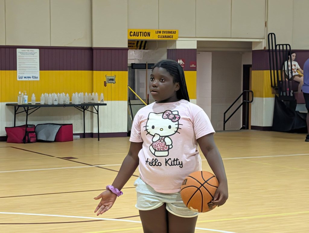 A girl wearing a Hello Kitty shirt holds a basketball in a gymnasium, with water bottles and bags on a table in the background.