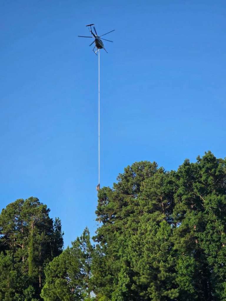 A helicopter carries a load suspended by a long cable above tall green trees, set against a clear blue sky.