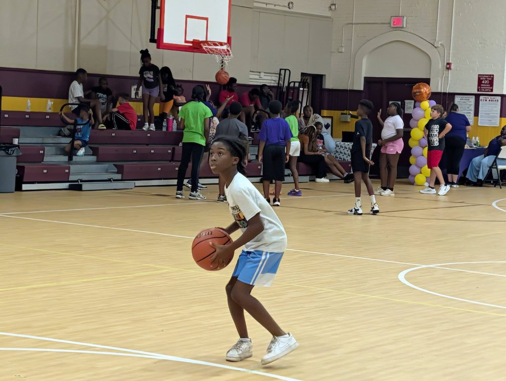 A young boy prepares to shoot a basketball in a busy gymnasium, with other children and adults in the background.