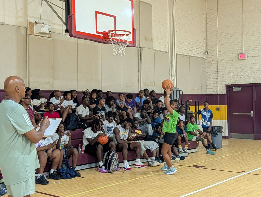 A woman prepares to shoot a basketball in a gym, while a coach observes and a group of young people sit watching on the bleachers.