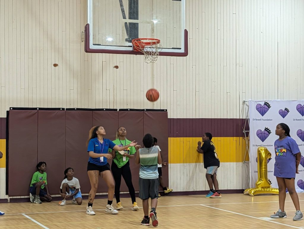 Kids playing basketball in a gym; some watching, others preparing to catch a rebound. DJ Rowell Foundation banner and gold balloon number one in the background.