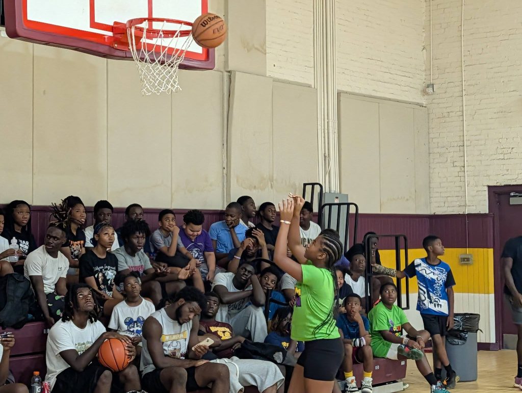 A girl shoots a basketball in a gym while a group of kids and teens watch from the bleachers.