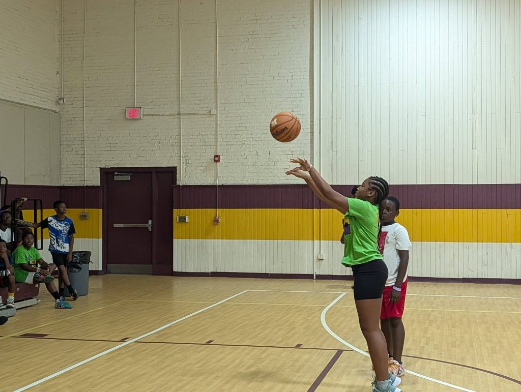 A girl in a green shirt shoots a basketball in an indoor gym while other kids watch from the bleachers.