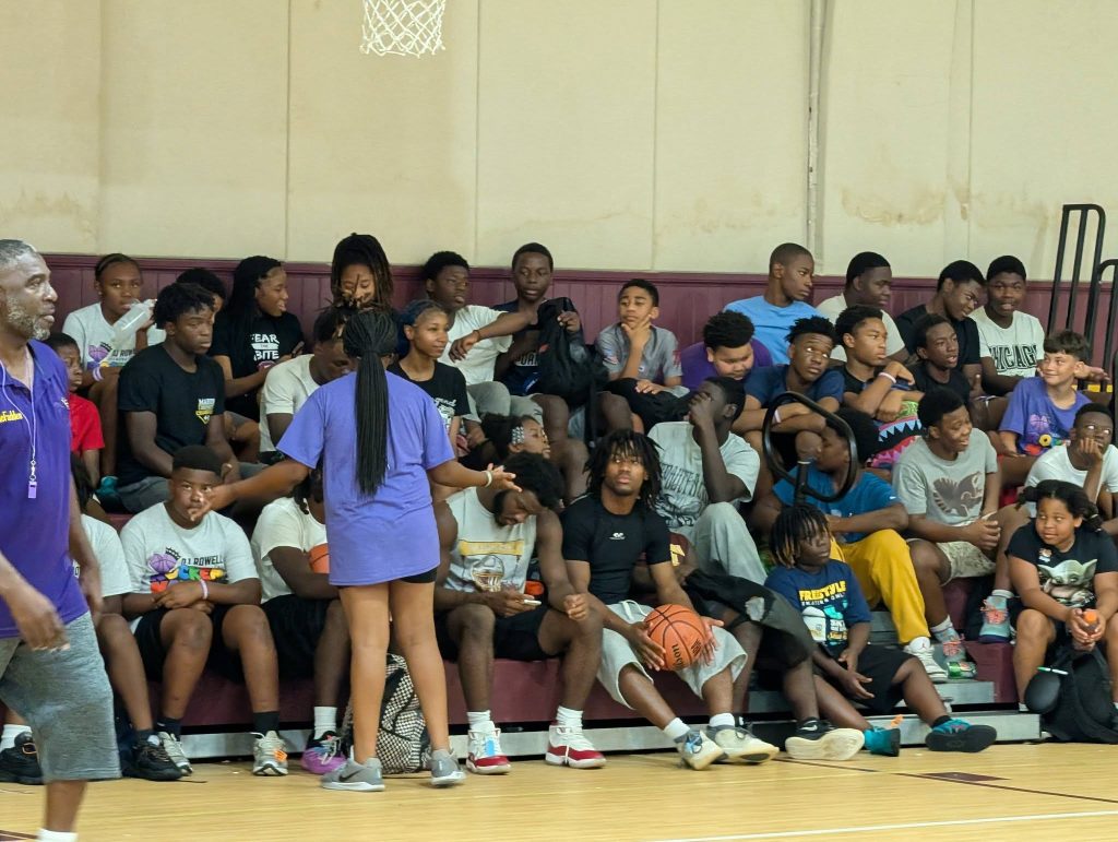 A group of teens and a coach sit on bleachers in a gym, watching something off-camera. One girl stands and talks to others, while one boy holds a basketball.