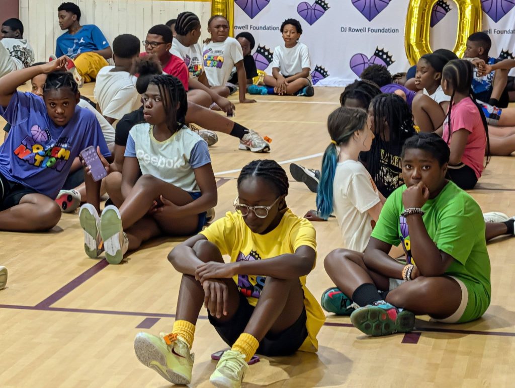 A group of children sit on a gym floor, some talking and others looking thoughtful, with a DJ Rowell Foundation banner in the background.