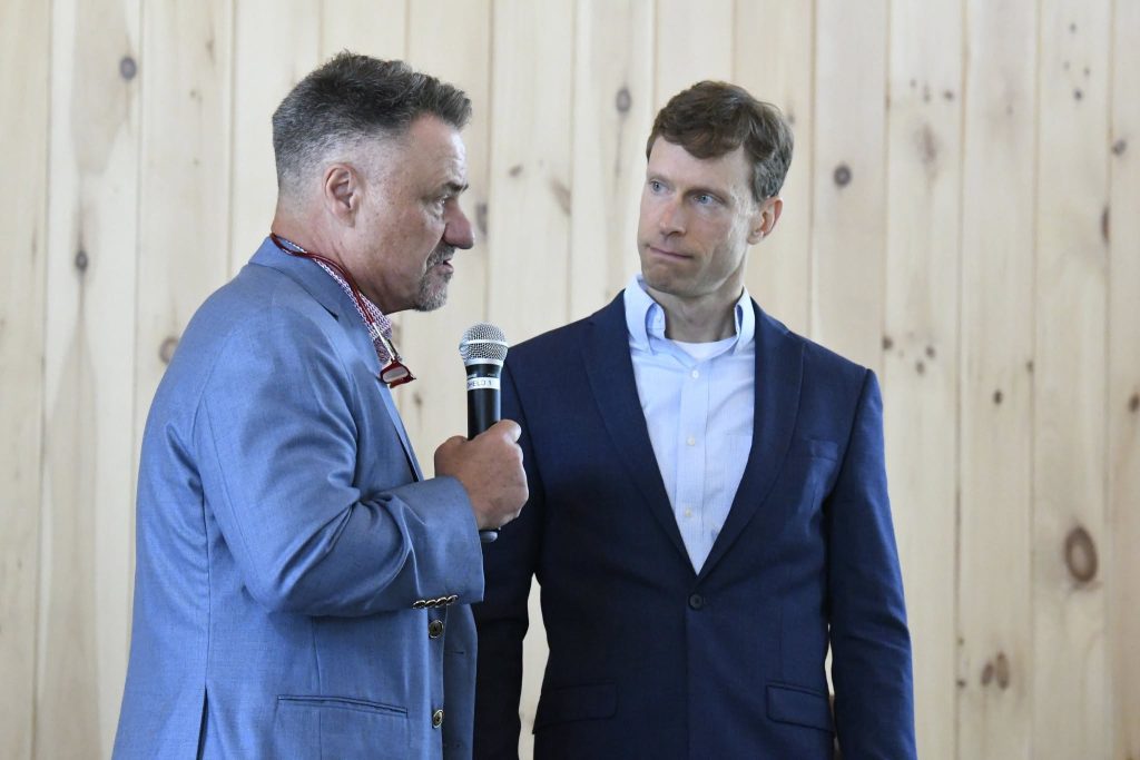Two men in suits standing indoors, one speaking into a microphone, the other attentively listening, with a wooden wall background.