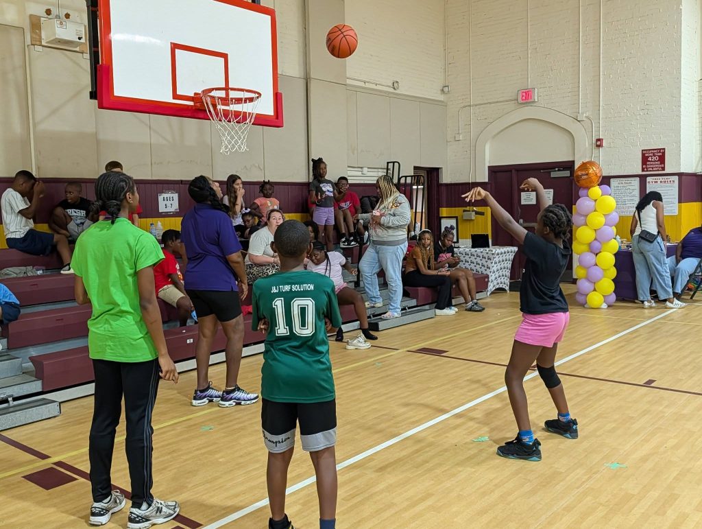 Children playing basketball in a gymnasium, with spectators sitting on bleachers in the background. A girl is shooting the ball toward the hoop.
