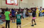 Children playing basketball in a gymnasium, with spectators sitting on bleachers in the background. A girl is shooting the ball toward the hoop.