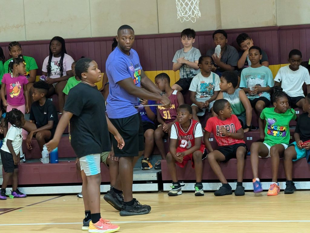 A group of children sit on gym bleachers while a man and a boy stand in front of them on a basketball court.