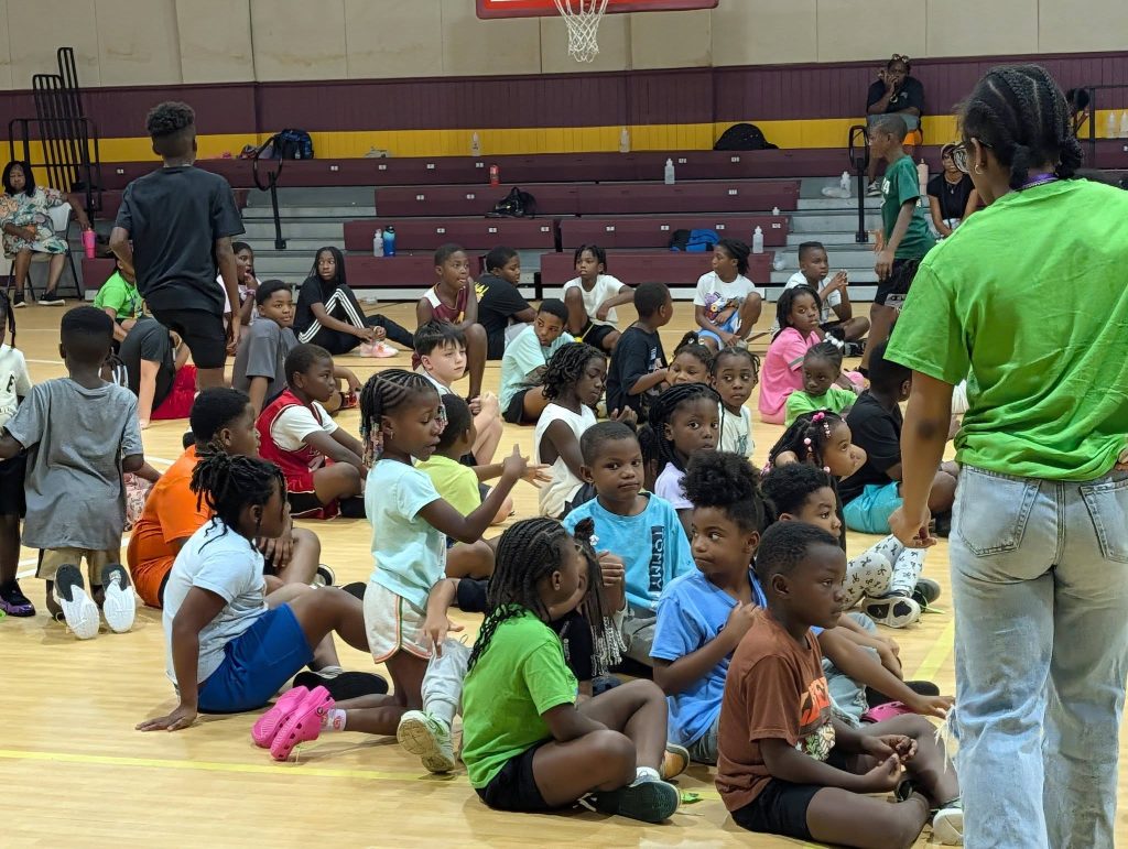 Children sit on a gym floor, listening to an adult with braided hair in a green shirt; a few other adults and kids are in the background.