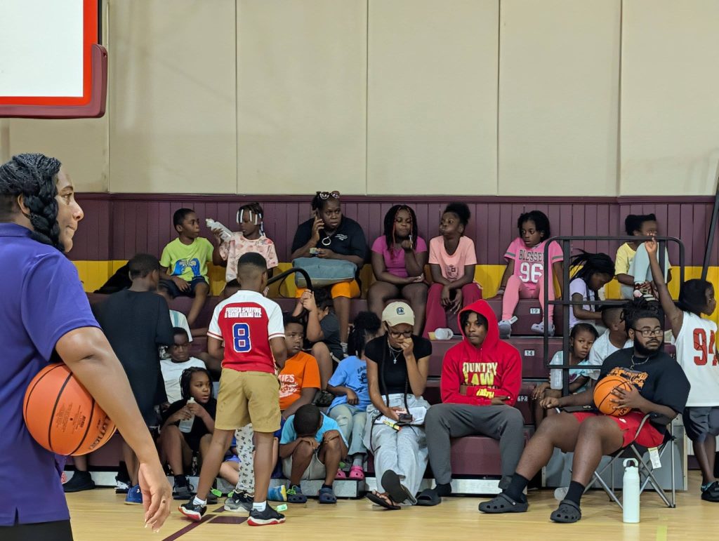 A group of children and adults sit on bleachers in a gym, some holding basketballs, while one woman stands holding a basketball on the court.