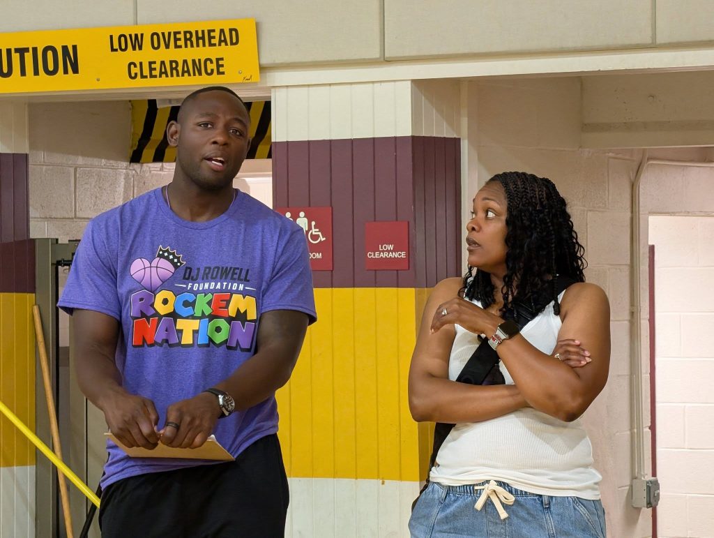 A man in a purple "Rockem Nation" shirt stands with a woman in a gym, under a yellow "Low Overhead Clearance" sign.