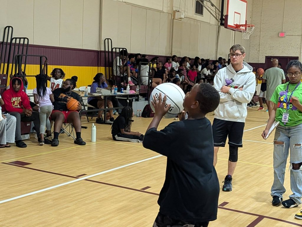 A young boy prepares to shoot a basketball in a busy indoor gym, while people watch from the bleachers and sidelines.