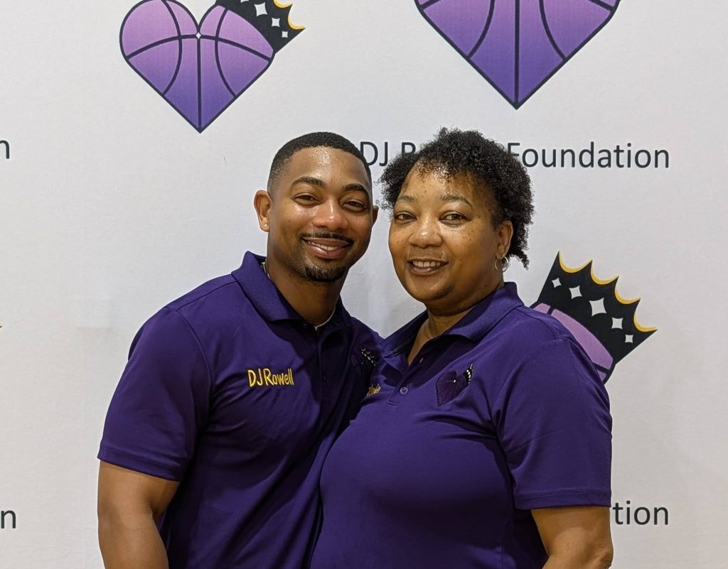 Two people wearing matching purple polo shirts stand smiling in front of a DJ Rowell Foundation banner with purple heart-shaped basketball logos.