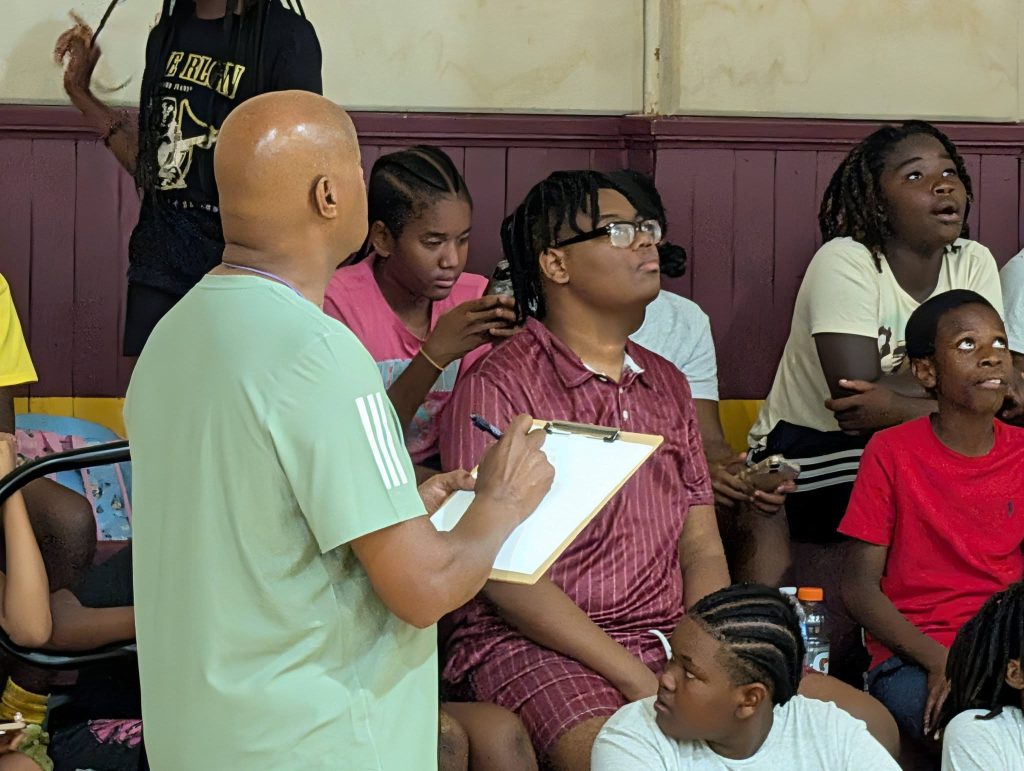A group of kids sitting in a gym, while an adult holding a clipboard stands in front, possibly observing or taking notes.