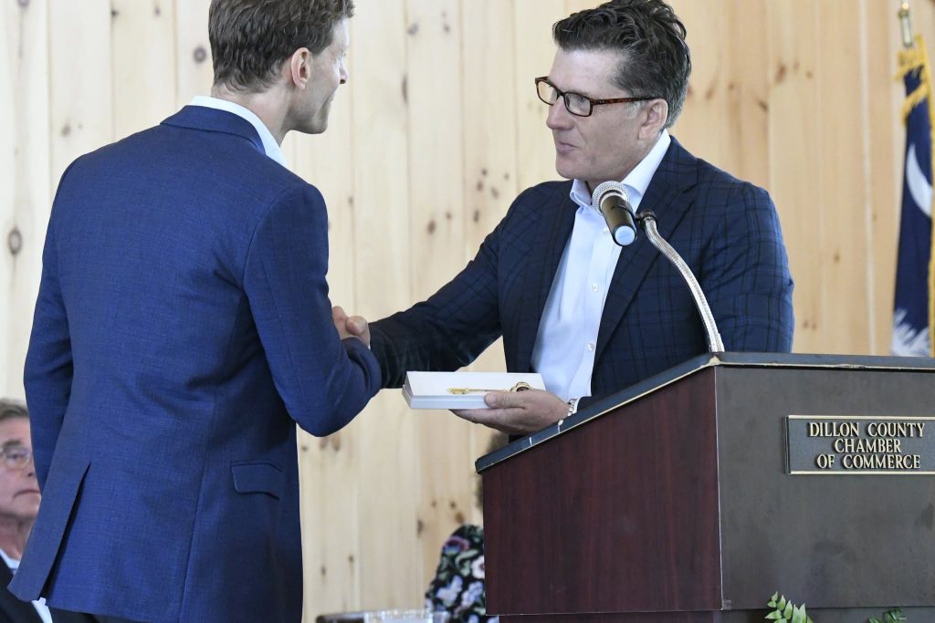 Two men in suits shake hands near a podium labeled "Dillon County Chamber of Commerce"; one is holding a ceremonial key in a box.